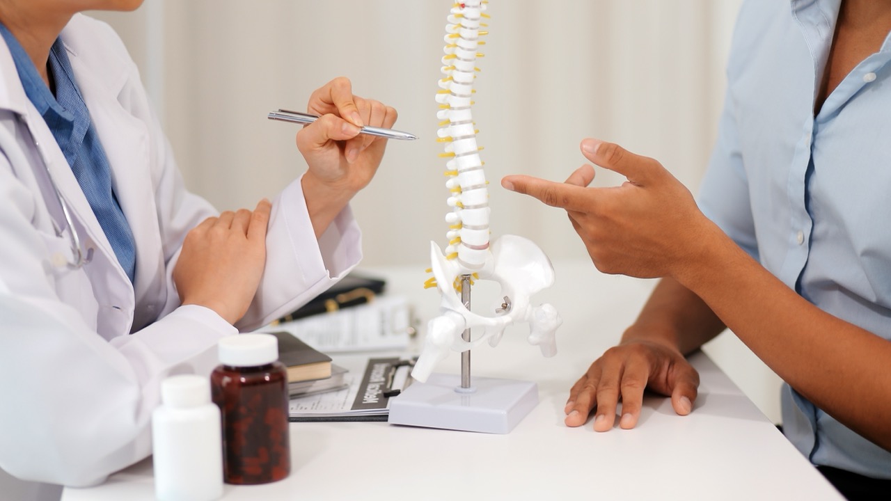 Doctor and patient discussing spinal health and osteoporosis while pointing to a model of the human spine during a medical consultation.
