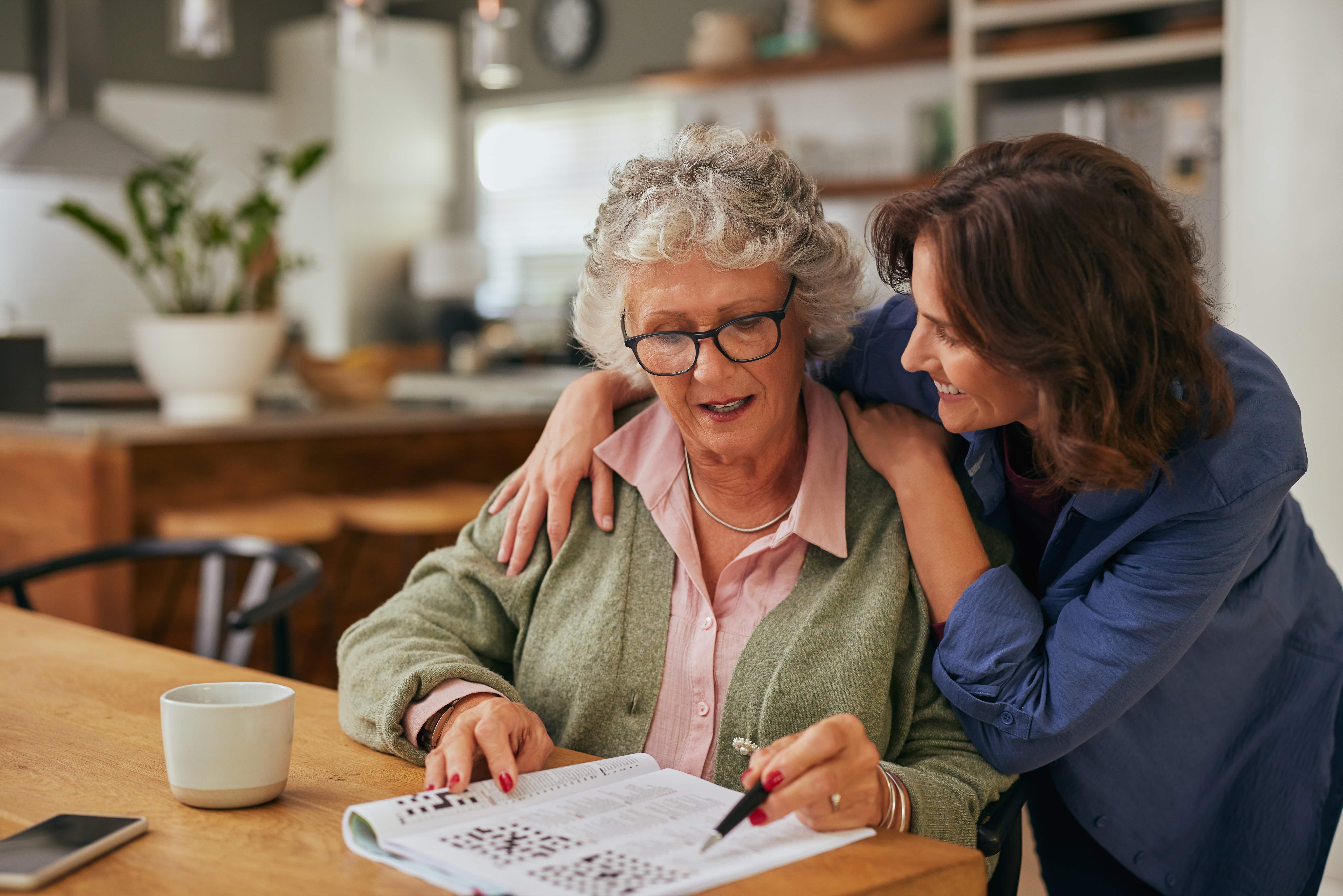 Smiling middle-aged woman embracing her elderly mother while they sit together at a kitchen table. The older woman, wearing glasses and a green cardigan, is working on a crossword puzzle, while a cup of coffee and a smartphone rest nearby.
